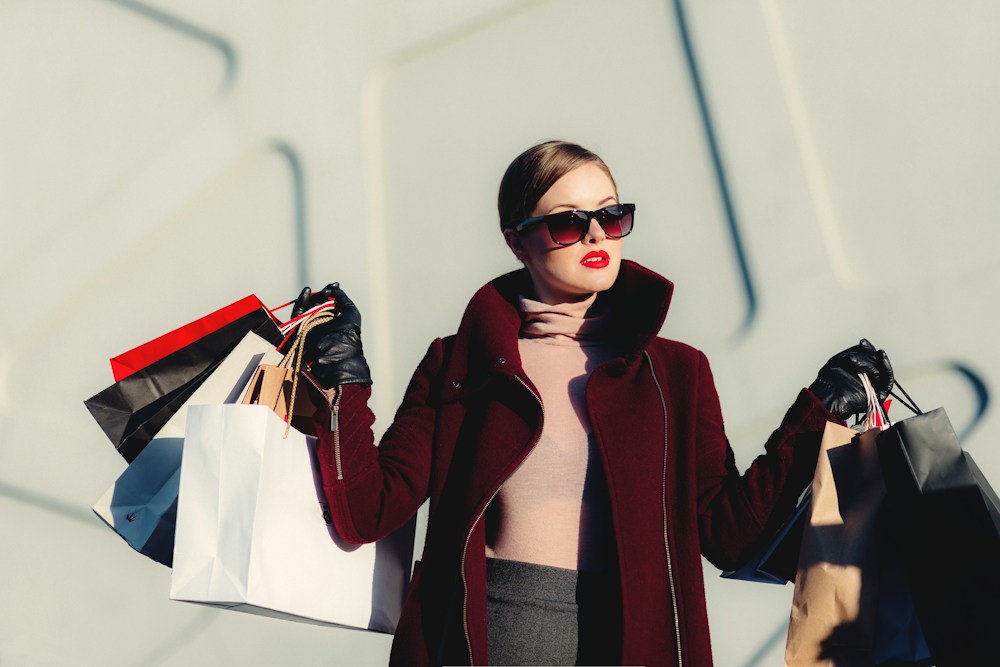 Woman in a burgundy coat and sunglasses holding shopping bags